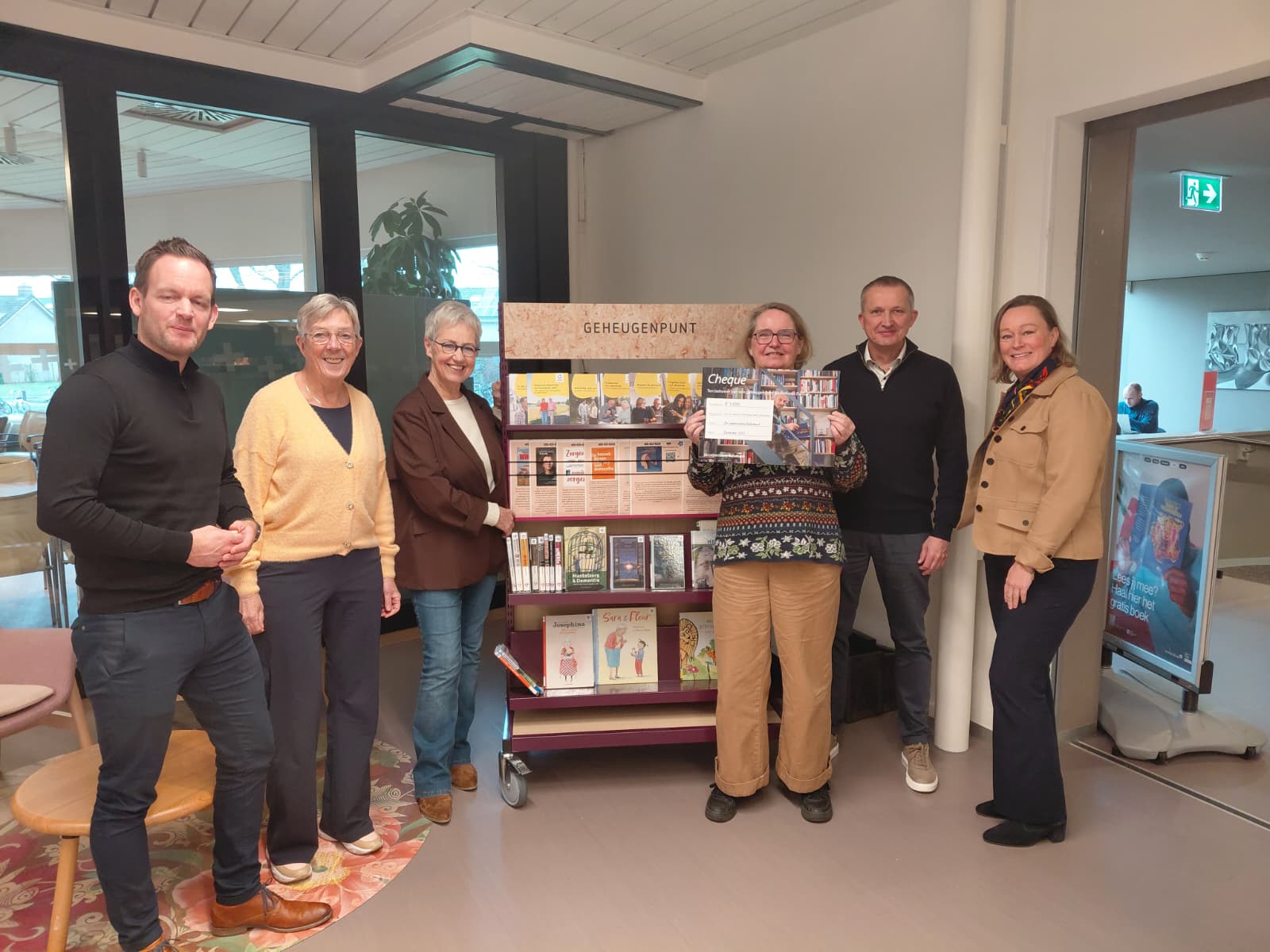 6 mensen staan naast een rek vol informatie over dementie en houden een cheque omhoog.