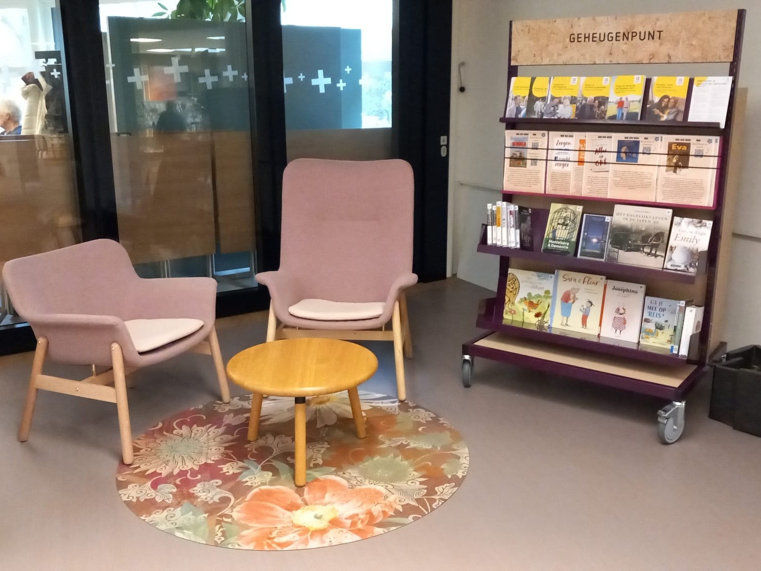 Rek met informatie over dementie, daarnaast staan twee stoelen en een tafeltje op een gebloemd vloerkleed.