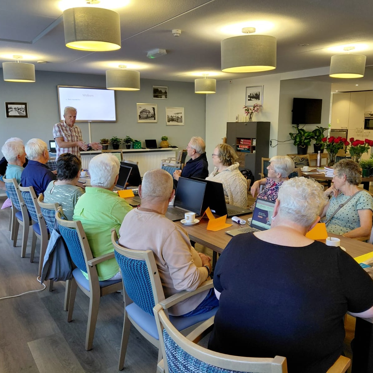 Groep deelnemers zitten aan een lange tafel met laptop voor zich en luisteren allemaal naar een vrijwilliger die wat uitlegt. 