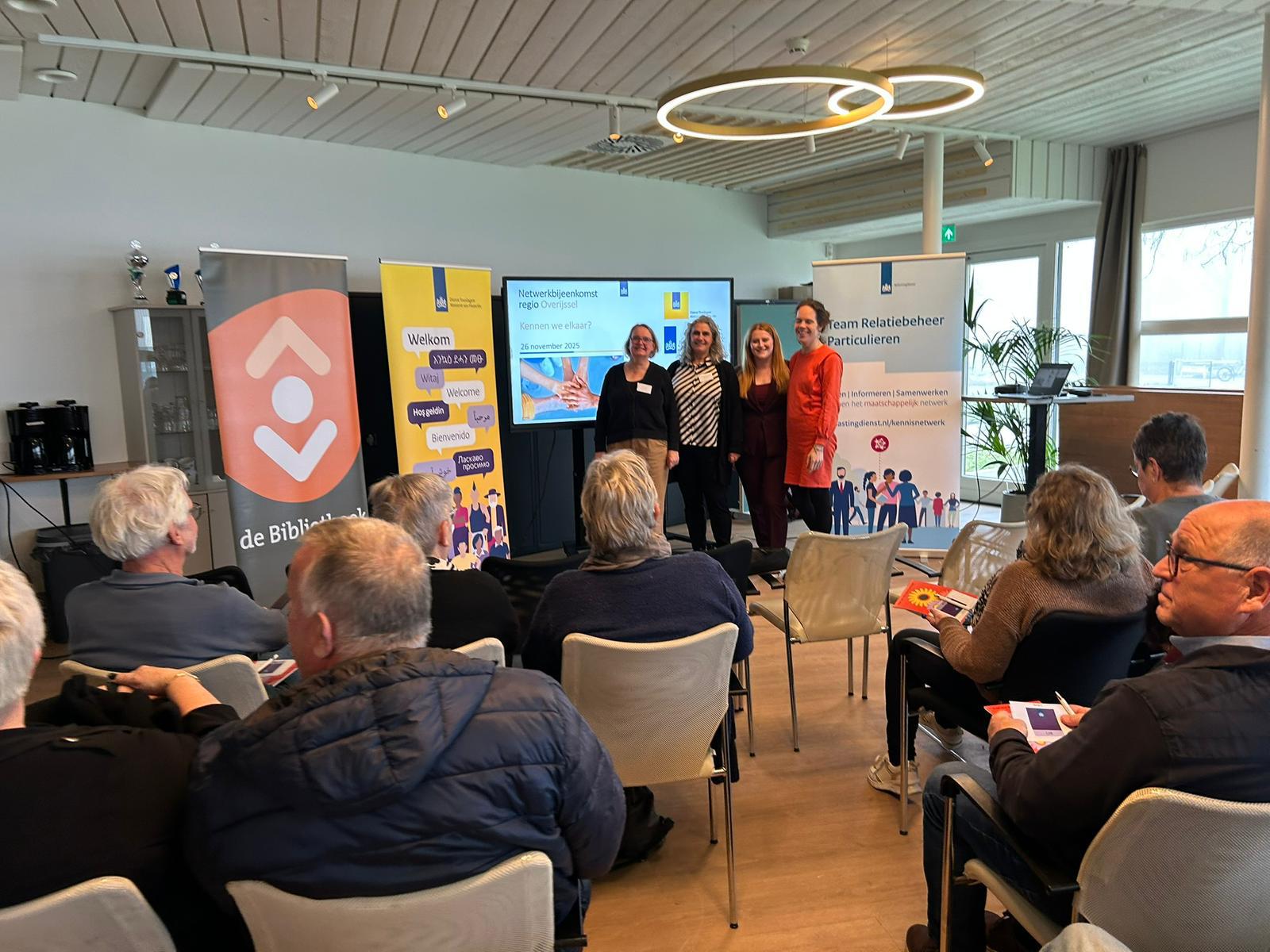 4 medewerkers van de belastingdienst en bibliotheek staan lachend naast een groot scherm, voor een groep deelnemers 