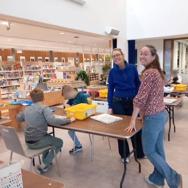 Twee medewerkers van de bibliotheek staan lachend bij een tafel terwijl kinderen druk bezig zijn met lego ontwerpen