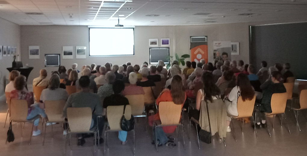 Grote groep mensen zit in een donkere zaal naar een scherm te kijken.